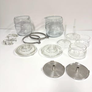 May include: A collection of clear glass and metal coffee maker parts, including glass carafes, lids, and metal filter components. The components are arranged on a white surface, showcasing the various pieces needed for brewing coffee.