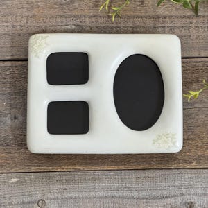 May include: A rectangular white picture frame with three openings: two squares and one oval. The frame has a floral design in the corners. The frame is on a wooden surface.