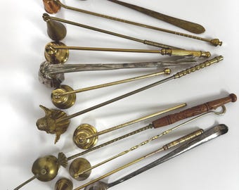 May include: A collection of various metal candle snuffers and long-handled tools. The set includes several brass, copper, and silver-toned items with diverse handle designs, including twisted metal and carved wood, arranged in a fan-like pattern.