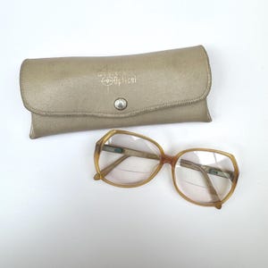 May include: Vintage eyeglasses with a light brown frame and clear lenses, next to a matching beige case. The case is embossed with "Western Optical" and a logo. The glasses have a classic, retro style.