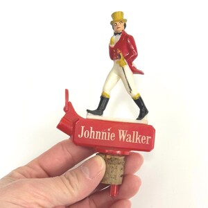 Puede incluir: Un tap&oacute;n de botella rojo con una figura en miniatura del Johnnie Walker Striding Man. La figura viste un abrigo rojo, pantalones blancos y un sombrero de copa amarillo. El tap&oacute;n tiene las palabras "Johnnie Walker" en blanco.