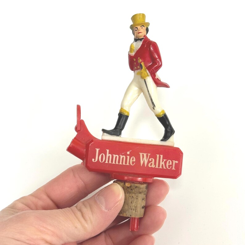 Puede incluir: Un tap&oacute;n de botella rojo con una figura en miniatura del Johnnie Walker Striding Man. La figura viste un abrigo rojo, pantalones blancos y un sombrero de copa amarillo. El tap&oacute;n tiene las palabras "Johnnie Walker" en blanco.