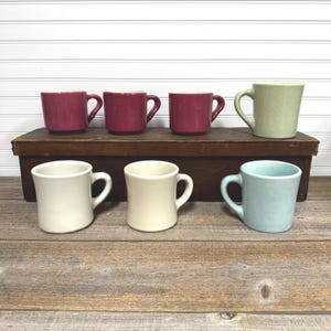 May include: A collection of ceramic mugs in various colors, including deep red, cream, light green, and speckled blue. The mugs are arranged on a wooden box and a wooden surface, showcasing their different hues and designs. The mugs have a classic, simple design.