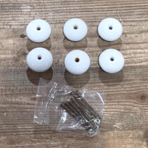 May include: Six white ceramic cabinet knobs with a glossy finish, each with a central hole. The knobs are arranged on a wooden surface, accompanied by a packet of screws and hardware, suggesting a set for furniture or cabinetry.
