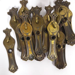 May include: A collection of antique brass drawer pulls. Each pull features an ornate design with a central circular element, a decorative top, and a curved handle. The pulls have a vintage, aged appearance with variations in color.