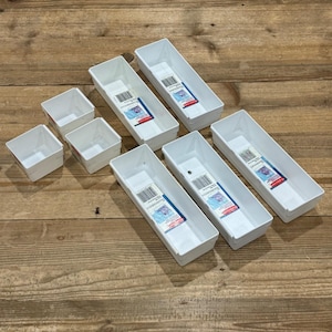 May include: A set of seven white plastic drawer organizers in various rectangular and square shapes. Each long rectangular bin features a product label with a barcode and blue and red branding text, while the smaller square bins are plain.
