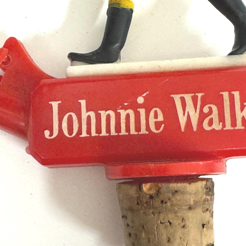 Puede incluir: Tap&oacute;n de botella rojo con el texto "Johnnie Walker" en blanco. El tap&oacute;n presenta una figura con botas negras y detalles amarillos. El corcho es visible en la parte inferior. Un objeto de colecci&oacute;n vintage.