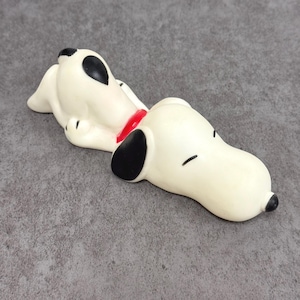 May include: A white Snoopy figurine in a relaxed pose, with black ears and spots, a red collar, and closed eyes. The figurine is lying down, with its head resting on its paws. The overall design is simple and cartoonish.