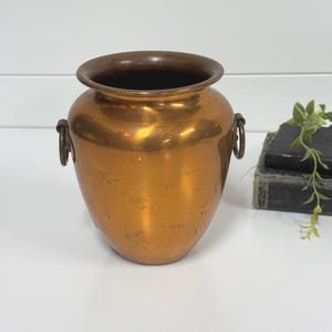 May include: A polished copper vessel with a wide, rounded opening and a tapered base. The container features two circular metal ring handles attached to opposite sides of the neck, showcasing a warm metallic finish with subtle surface variations.