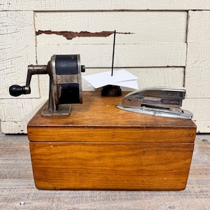 May include: A wooden box with a pencil sharpener, a stapler, and a piece of paper on top. The box is brown and has a simple design.