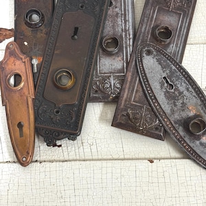 May include: A collection of antique door hardware, including ornate bronze and copper keyhole covers and doorknob backplates. The pieces show signs of age and wear, with a distressed, vintage aesthetic. The hardware is arranged on a white, cracked paint surface.