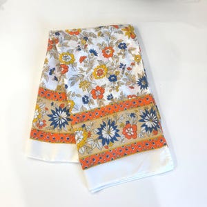 May include: A folded white fabric featuring a vibrant floral pattern with orange, yellow, and dark blue blossoms and green leaves. A horizontal border band displays a repeating geometric orange design and large blue flowers on a tan background.