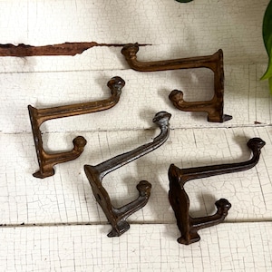 May include: Five antique-style metal wall hooks in various shades of brown and gray. Each hook has a curved design with a rounded knob at the top and a smaller knob below. The hooks are arranged on a distressed white wooden surface.