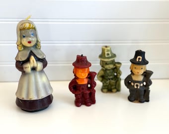 May include: A collection of vintage Thanksgiving-themed candles. The set includes a pilgrim girl, a pilgrim man, and two other figures. The candles are in various colors, including white, burgundy, orange, and green. The candles are on a white surface.