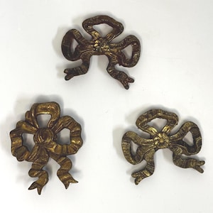 May include: Three antique-style, gold-colored bow ornaments. Each bow has a detailed, textured design with multiple loops and tails. The bows are arranged on a white background, showcasing their intricate details and vintage aesthetic.