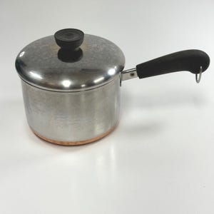 May include: A stainless steel saucepan with a copper-colored base, a matching stainless steel lid with a black knob, and a black handle featuring a small metal hanging loop at the end. The pot has a cylindrical body and a polished finish.