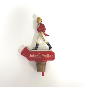 Puede incluir: Un tap&oacute;n de botella rojo con el logotipo de Johnnie Walker. El tap&oacute;n tiene una base de corcho y una parte superior de pl&aacute;stico rojo con el nombre de la marca y una figura del Striding Man con abrigo rojo, pantalones blancos y botas negras.