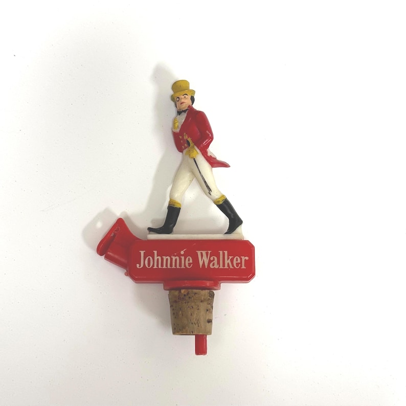 Puede incluir: Un tap&oacute;n de botella rojo con el logotipo de Johnnie Walker. El tap&oacute;n tiene una base de corcho y una parte superior de pl&aacute;stico rojo con el nombre de la marca y una figura del Striding Man con abrigo rojo, pantalones blancos y botas negras.