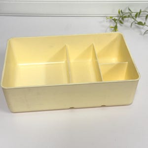 May include: A rectangular, pale yellow plastic organizer with four compartments. The organizer is designed to hold small items and has a simple, functional design. The compartments are divided by vertical dividers.