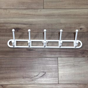 May include: A white metal coat rack with five hooks. The hooks are evenly spaced along a rectangular bar with rounded ends. Each hook has a ball-shaped top. The rack is mounted on a wooden surface.
