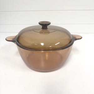 May include: A round, amber-colored glass casserole dish with a matching transparent lid. The dish features two integrated side handles and a central knob on the lid. The glass is translucent, allowing light to pass through the brown-tinted material.