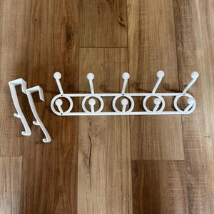 May include: White metal over-the-door hook rack with six rounded hooks and a decorative design. Includes two additional over-the-door hooks. The hooks are designed for hanging clothes, towels, or other items. The rack is made of metal and is painted white.