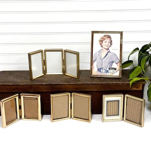 May include: A collection of vintage gold-toned picture frames. The frames vary in size and style, including single, double, and triple folding designs. One frame contains a vintage advertisement. The frames are displayed on a dark wooden surface.