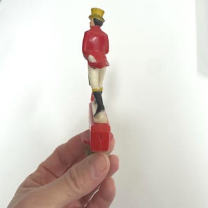 Puede incluir: Figura vintage de un hombre con abrigo rojo, sombrero de copa amarillo y botas negras. La figura est&aacute; de pie sobre una base roja. La figura es un objeto decorativo.