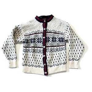 May include: A cream-colored cardigan sweater with a detailed pattern. The sweater features a navy blue and burgundy trim, with a snowflake and geometric design. It has a button-up front and long sleeves.
