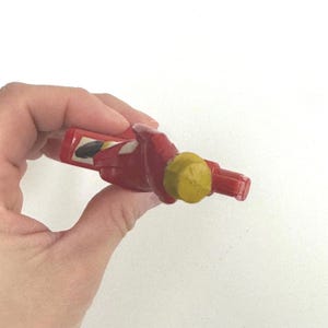 Puede incluir: Una botella roja con tap&oacute;n amarillo se sostiene en una mano. La botella tiene una peque&ntilde;a etiqueta rectangular con un dise&ntilde;o en blanco y negro. La botella es de pl&aacute;stico duro. Estilo vintage.