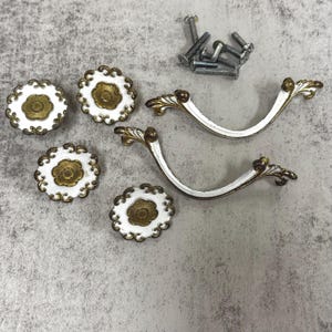 May include: A set of white and gold cabinet hardware, including four round knobs and two pull handles. The knobs have a floral design with a gold center and scalloped edges. The handles are white with gold accents and decorative flourishes. A small pile of screws is also present.