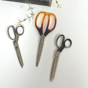 May include: Three pairs of scissors are arranged on a white surface. One pair has a weathered metal finish, one features translucent amber-colored plastic handles, and the third pair has black painted handles with visible signs of wear.