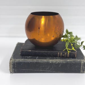 May include: A spherical, copper-colored bowl with a dark interior sits atop two antique, dark-covered books. The bowl reflects light, showcasing its metallic finish. A sprig of green foliage adds a natural accent to the composition.