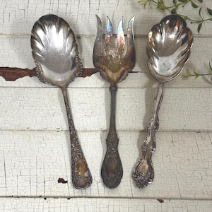 May include: Three antique silver serving utensils: a spoon, a fork, and another spoon. The spoon on the left has a large, scalloped bowl and ornate handle. The fork has four tines. The spoon on the right has a smaller, scalloped bowl.
