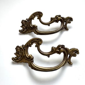 May include: Two antique brass drawer pulls with ornate, scrolling designs. The pulls have a curved handle and decorative floral accents. The brass has a warm, aged patina, suggesting vintage or antique origins. These pulls are suitable for furniture restoration or adding a vintage touch.