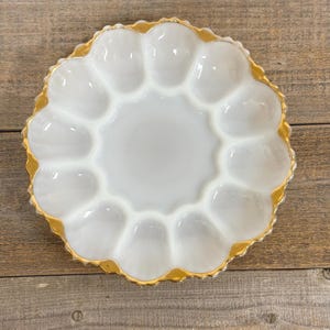 May include: White, scalloped egg serving dish with a gold rim. The dish has 12 individual egg compartments surrounding a central serving area. The dish is made of a glossy, opaque material and sits on a wooden surface.