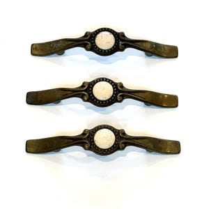 May include: Three antique-style cabinet pulls with a bronze finish. Each pull features a central, speckled, off-white ceramic disc surrounded by a decorative border. The pulls are curved and have a classic, vintage aesthetic.