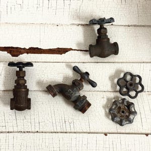 May include: An assortment of vintage metal faucet parts, including valves and handles, in various sizes and designs. The pieces have a weathered, aged appearance with visible rust and wear, suggesting they are reclaimed or antique items. The background is a white, distressed wood surface.