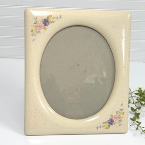 May include: Cream-colored ceramic picture frame with an oval opening. The frame has a crackle finish and floral accents in pink, purple, and yellow at the corners. The frame is rectangular with rounded corners.