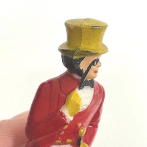 Puede incluir: Figura vintage de un hombre con abrigo rojo, sombrero de copa amarillo y guantes amarillos, sosteniendo un mon&oacute;culo. La figura tiene una pajarita negra y est&aacute; pintada con detalles. Es un objeto de colecci&oacute;n.