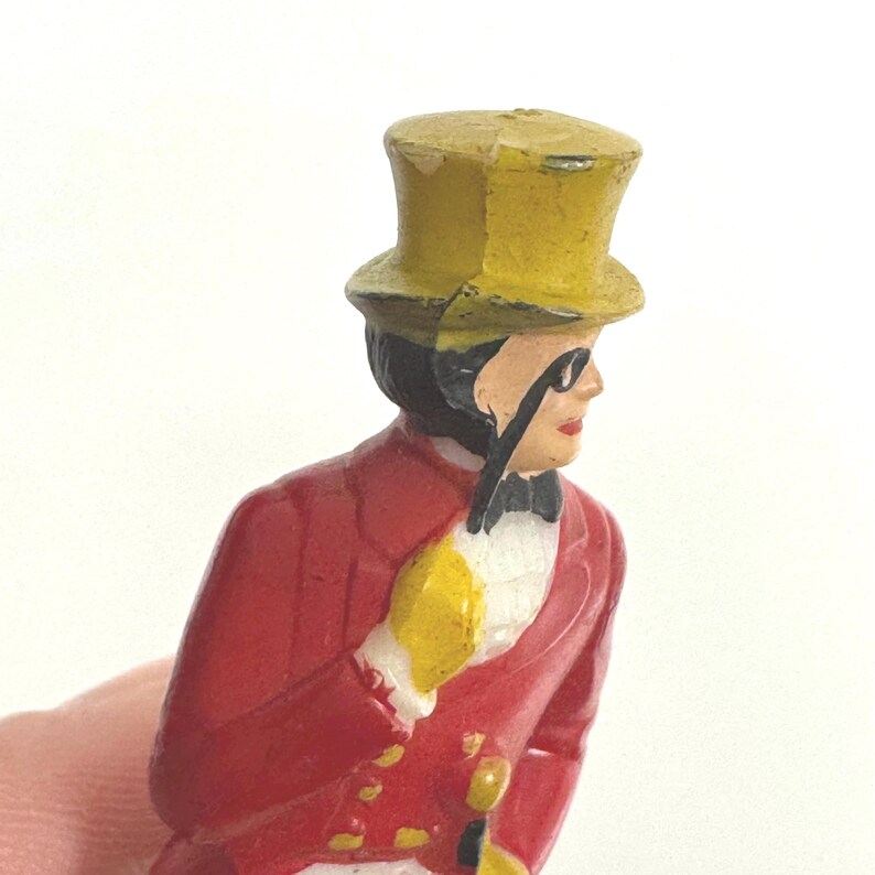 Puede incluir: Figura vintage de un hombre con abrigo rojo, sombrero de copa amarillo y guantes amarillos, sosteniendo un mon&oacute;culo. La figura tiene una pajarita negra y est&aacute; pintada con detalles. Es un objeto de colecci&oacute;n.
