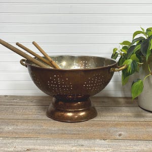 May include: A vintage copper colander with a pedestal base and two handles. The colander has numerous small holes and contains three wooden stirring spoons. A green plant in a white pot is visible in the background.