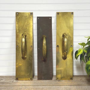 May include: Three vintage brass door pull handles. Two are a bright gold color, and one is a dark bronze. Each handle has a rectangular backplate with a curved handle. The handles are mounted on a white-paneled wall.