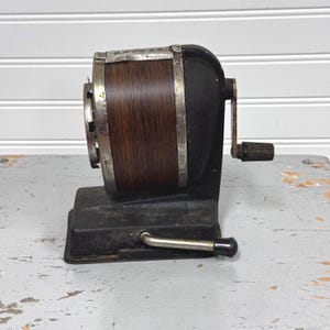 May include: Vintage black and brown pencil sharpener with a wooden barrel and silver accents. The sharpener has a crank handle and a base with a lever. The word "BOSTON" is visible on the top of the sharpener.