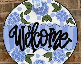 Könnte beinhalten: Ein rundes Türschild aus Holz mit weißem Hintergrund, blauen Punkten und blauen Hortensienblüten mit grünen Blättern. Ein horizontales blau gestreiftes Band verläuft durch die Mitte mit dem Wort Welcome in schwarzer Schreibschrift.
