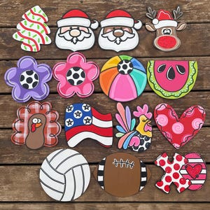 May include: A collection of fourteen decorative wooden cutouts featuring various shapes including a Christmas tree, Santa Claus faces, a reindeer, flowers, a beach ball, a watermelon slice, a turkey, an American flag, a rooster, a heart, a volleyball, a football, and an XOXO sign.