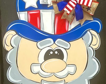 May include: A wooden door hanger featuring a cartoon Uncle Sam with a blue hat, red and white stripes, and a white beard. The hat is decorated with a red, white, and blue bow.