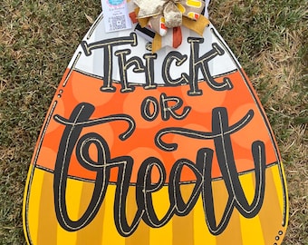 May include: A large, wooden candy corn-shaped sign with the words "Trick or Treat" in black script. The sign is painted in the traditional candy corn colors of white, orange, and yellow. A decorative bow with candy corn accents and a tag are attached to the top.