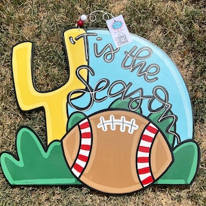 May include: A wooden door hanger with a football, green grass, and a yellow goal post. The text "Tis the Season" is written in black cursive on a blue background.