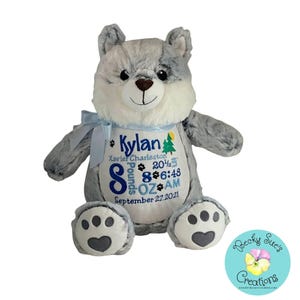 May include: A plush gray and white wolf toy with a light blue ribbon around its neck. The white belly features embroidered text: Kylan, Xavier Charleston, 8 Pounds, 8 OZ, 20 1/2, 6:48 AM, September 27, 2021, with paw prints, a tree, and a sun.